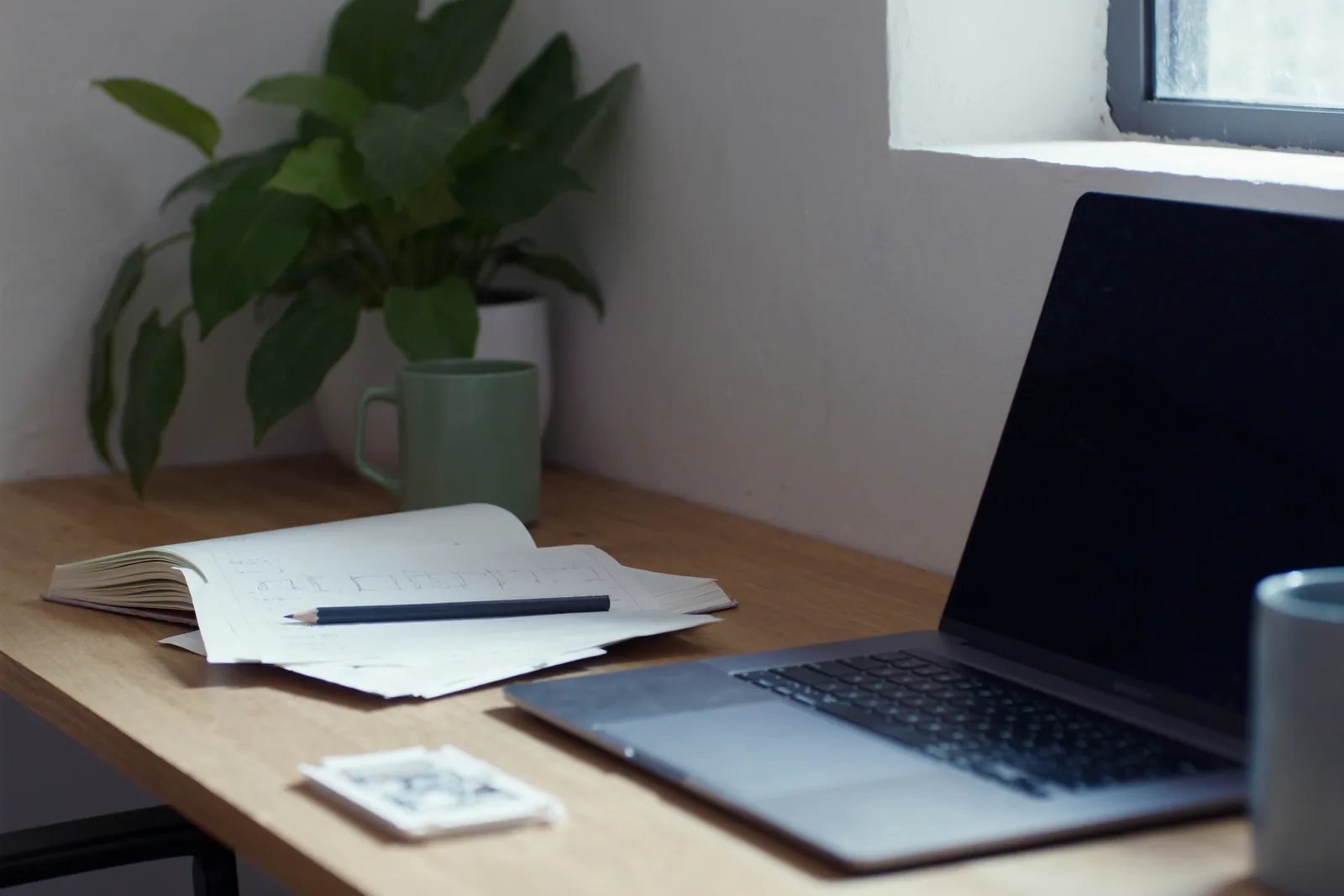 A designer's desk by a window, a laptop with its screen dark and a notebook with a pencil sketch of a page